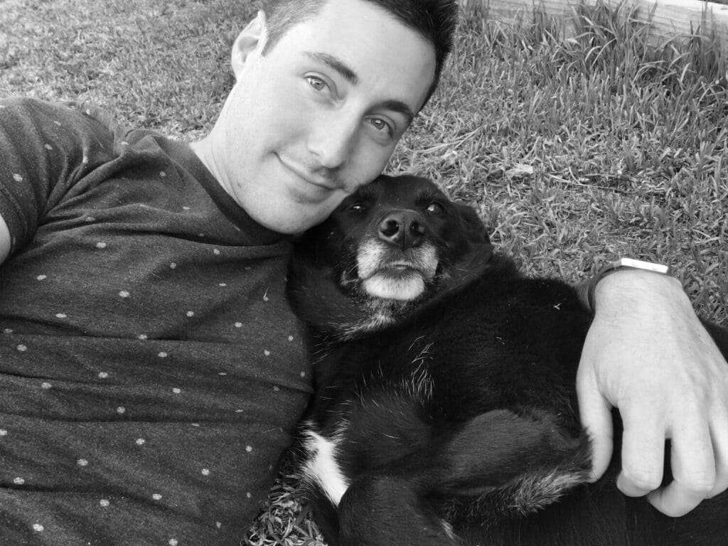 Black & white image of Ben lying on some grass cuddling a black & white dog under his left arm.