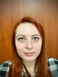 Headshot of Gabriela, standing against a wooden background. She has a chequered shirt on.