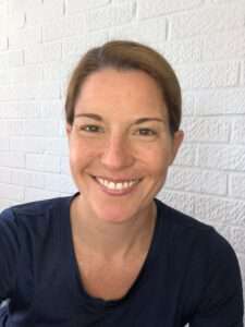 Image of Sandra wearing a navy top against a white brick wall. She is smiling.
